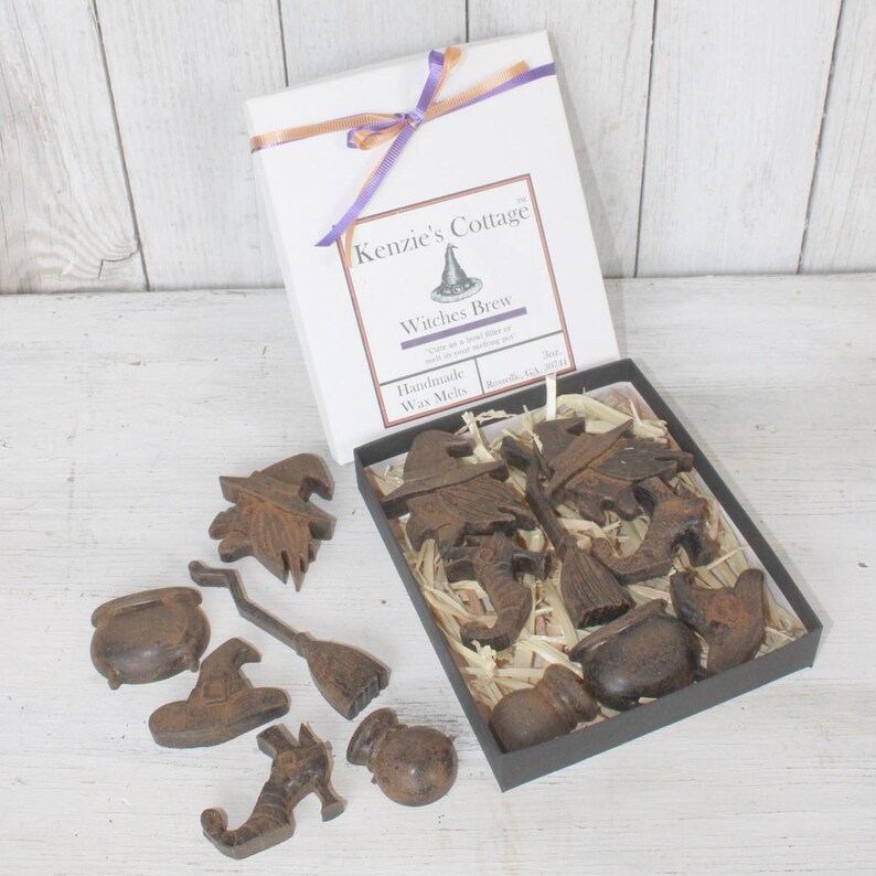 Op de afbeelding: Een set handgemaakte wax melts in de vorm van Halloween-objecten, waaronder heksen, ketels, bezems en vleermuizen. De wax melts zijn verpakt in een zwarte doos met een wit deksel waarop "Kenzie's Cottage" en "Witches Brew" staat.