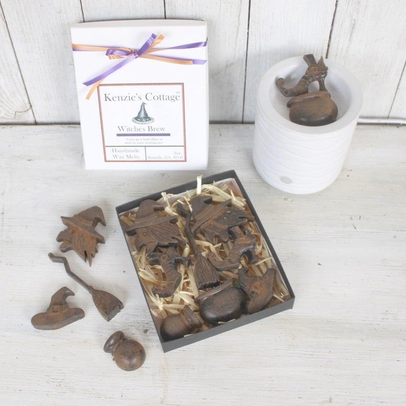 Op de afbeelding: Een doos met handgemaakte wax melts met een heksenthema. De doos is gelabeld met "Kenzie's Cottage" en "Witches Brew". De wax melts zijn gevormd als heksen, bezems, ketels en hoeden.