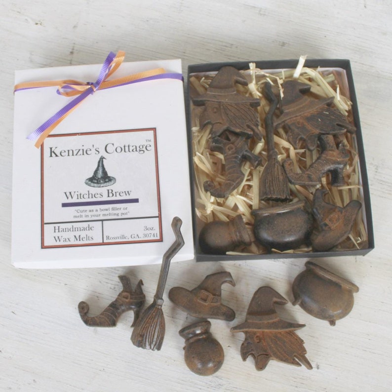 Op de afbeelding: Een doos met handgemaakte wax melts in de vorm van heksen, bezems, ketels en hoeden. De doos is wit met een paars lint en de tekst "Kenzie's Cottage" en "Witches Brew".