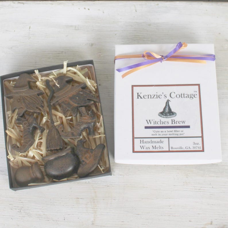 Op de afbeelding: Een witte doos met een paars en oranje lint eromheen gebonden. De doos heeft een label met de tekst "Kenzie's Cottage" en "Witches Brew Handmade Wax Melts". De doos bevat een set kleine, zwarte metalen wax melts in de vorm van heksen, bezems en ketels.