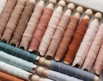 Può includere: Assortimento di bobine di nastro di seta tinti a mano in varie tonalità di marrone, rosa, blu e rosso. Il nastro è avvolto attorno a perni di legno.