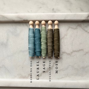 May include: Five spools of silk ribbon wrapped around wooden dowels. The ribbon colors are Robbins Egg blue, Laurel green, Sage green, Eucalyptus green, and Moss green. The colors are labeled below each spool.