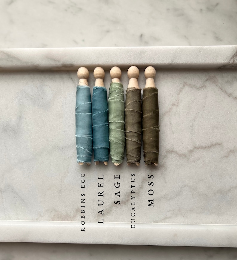 May include: Five spools of silk ribbon wrapped around wooden dowels. The ribbon colors are Robbins Egg blue, Laurel green, Sage green, Eucalyptus green, and Moss green. The colors are labeled below each spool.