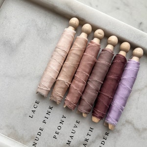 May include: Six spools of silk ribbon in various shades of pink, brown, and purple. The spools are labeled "Lace", "Nude Pink", "Peony", "Mauve", "Earth", and "Lavender".