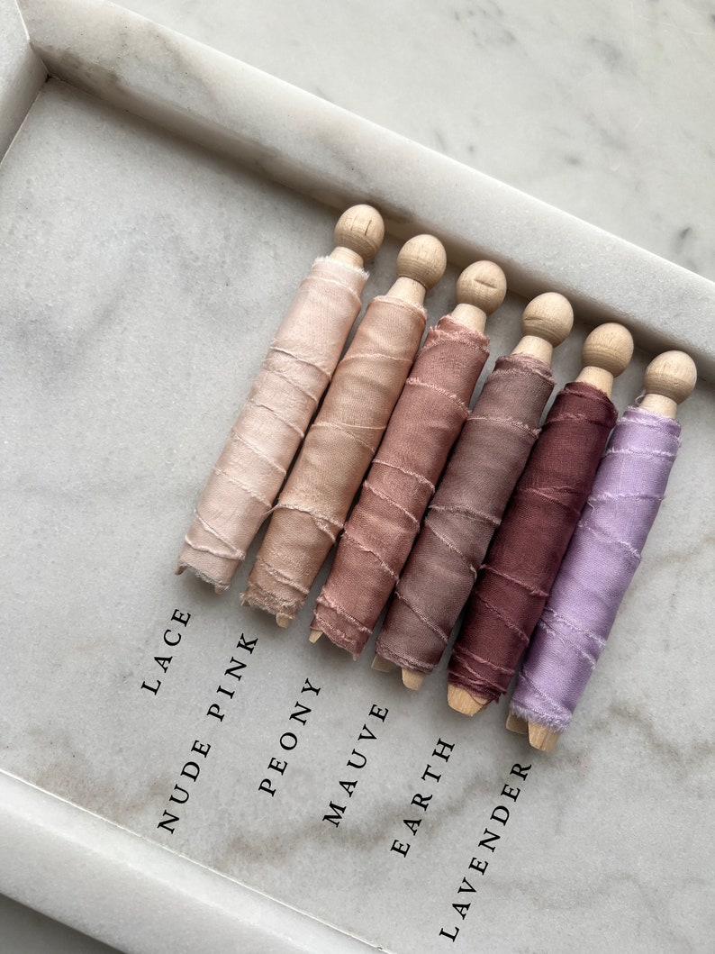 May include: Six spools of silk ribbon in various shades of pink, brown, and purple. The spools are labeled "Lace", "Nude Pink", "Peony", "Mauve", "Earth", and "Lavender".