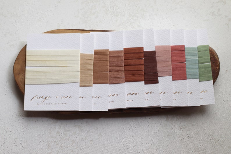 May include: A color palette of silk ribbons in various shades of beige, brown, and muted colors, displayed on white cards. The cards are arranged on a wooden tray. The text "fringe + rose" and "HAND-DYED SILK RIBBON" is visible.