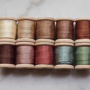 May include: A collection of ten wooden spools, each wound with a different shade of silk thread. Colors range from cream and gold to various shades of brown, red, and green. The spools are arranged in two rows of five.