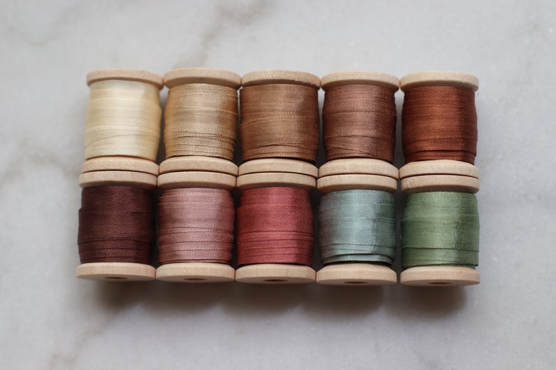 May include: A collection of ten wooden spools, each wound with a different shade of silk thread. Colors range from cream and gold to various shades of brown, red, and green. The spools are arranged in two rows of five.