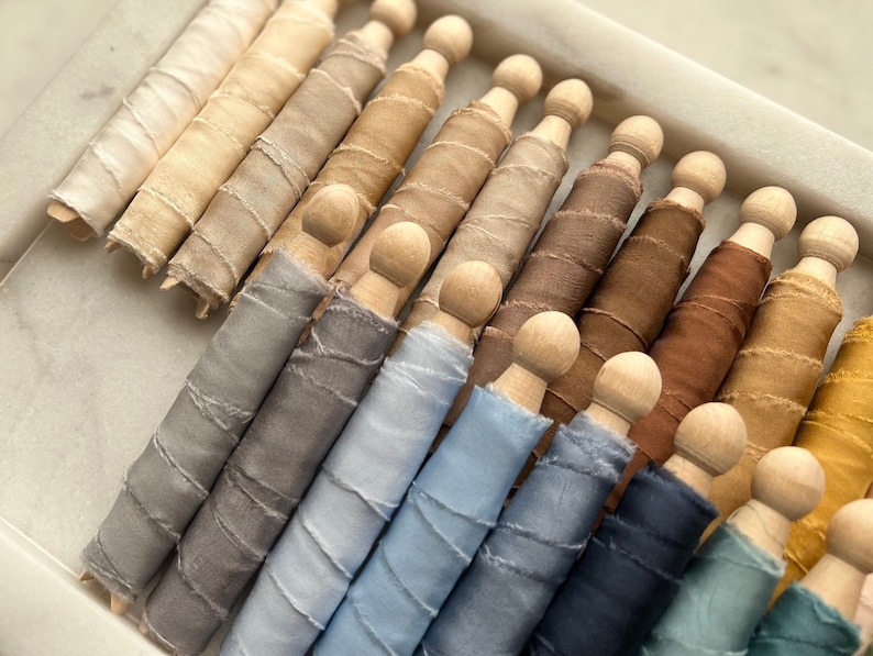 May include: A collection of 20 spools of silk ribbon in various shades of brown, gray, blue, and yellow. The ribbon is wound around wooden spools.