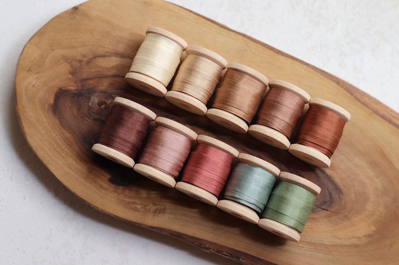 May include: A collection of ten wooden spools of thread in various earth-tone colors, including beige, brown, and muted greens and pinks. The spools are arranged on a wooden, oval-shaped tray.