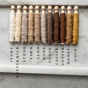 May include: Ten spools of silk ribbon wound around wooden dowels, arranged in a row from left to right. The colors are ivory, buttermilk, stone, fawn, sand, bone, taupe, coffee, copper, honey, and gold.