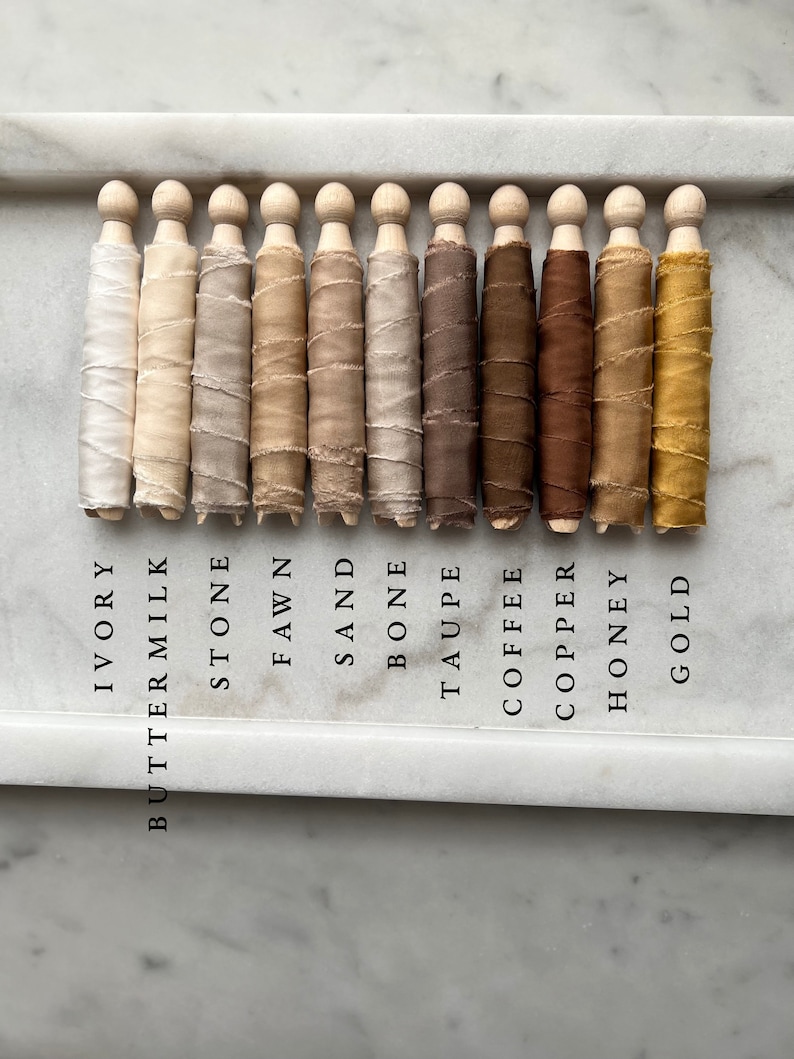May include: Ten spools of silk ribbon wound around wooden dowels, arranged in a row from left to right. The colors are ivory, buttermilk, stone, fawn, sand, bone, taupe, coffee, copper, honey, and gold.