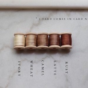 May include: Five wooden spools of thread in varying shades of neutral colors: ivory, wheat, camel, latte, and rust. The text below each spool identifies the color. The text at the top says "1 yard comes in card not spool."