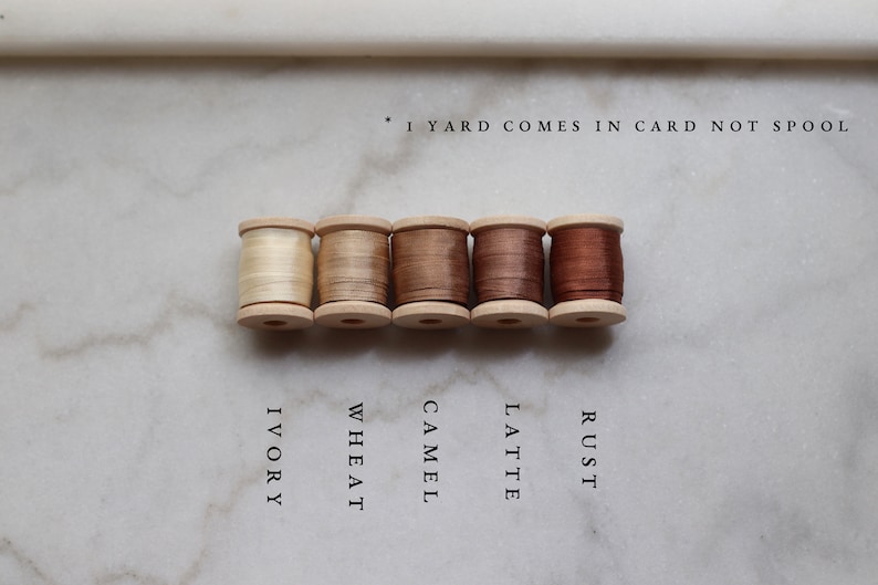 May include: Five wooden spools of thread in varying shades of neutral colors: ivory, wheat, camel, latte, and rust. The text below each spool identifies the color. The text at the top says "1 yard comes in card not spool."