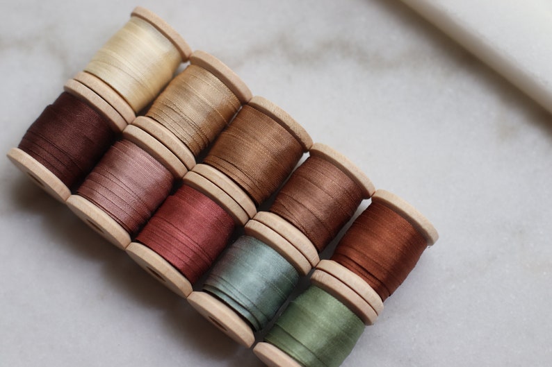 May include: A collection of wooden spools filled with various shades of thread, including beige, brown, pink, and green. The spools are arranged diagonally, showcasing the different colors and textures of the thread. Ideal for sewing and crafting projects.