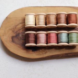 May include: A collection of ten wooden spools of silk thread in various earth-tone colors, including cream, beige, brown, and green. The spools are arranged on a natural wood serving board. The threads appear to be approximately 1 inch in diameter.