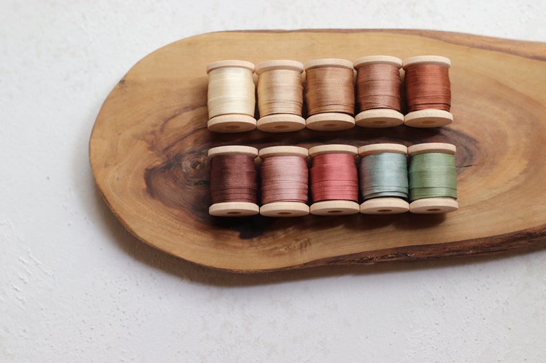 May include: A collection of ten wooden spools of silk thread in various earth-tone colors, including cream, beige, brown, and green. The spools are arranged on a natural wood serving board. The threads appear to be approximately 1 inch in diameter.