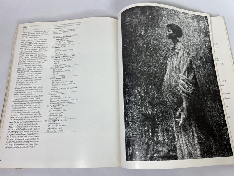 May include: An open art book featuring a biography of Charles White on the left page and a full-page black and white illustration of a person in a long, textured robe on the right page. The text includes detailed lists of artworks and exhibitions.