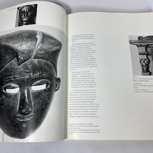May include: An open book page featuring a black and white photograph of a dark, carved wooden mask with a flat top and almond-shaped eyes. The text on the page discusses historical architecture and craftsmanship, including references to ironwork and slave labor.