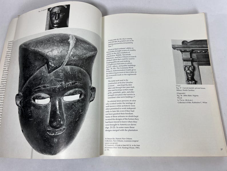 May include: An open book page featuring a black and white photograph of a dark, carved wooden mask with a flat top and almond-shaped eyes. The text on the page discusses historical architecture and craftsmanship, including references to ironwork and slave labor.