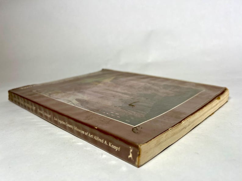 May include: A softcover book with a brown cover featuring a rectangular image on the front. The spine and cover display the text Los Angeles County Museum of Art and Alfred A. Knopf. The book is positioned at an angle, showing the front cover.