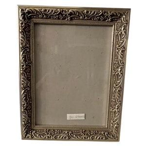 May include: A gold-toned picture frame with an ornate, detailed design. The frame has a rectangular shape and a smooth, polished finish.