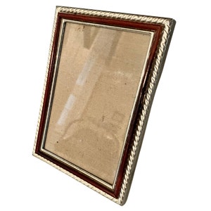 Puede incluir: Un marco de fotos rectangular con un borde plateado en forma de cuerda. El marco tiene un borde interior marr&oacute;n oscuro y un cristal transparente. El marco est&aacute; vac&iacute;o, listo para mostrar una foto o una obra de arte.