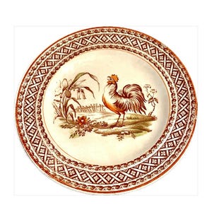 May include: A round, cream-colored plate with a brown rooster illustration in the center. The rooster is standing in front of a fence and flowers. The plate has a decorative brown border with a repeating geometric pattern.