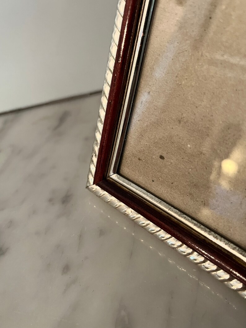 Puede incluir: Primer plano de un marco de fotos rectangular con un borde plateado en forma de cuerda y un marco interior marr&oacute;n oscuro. El marco contiene una imagen de color claro y se encuentra sobre una superficie de m&aacute;rmol blanco.