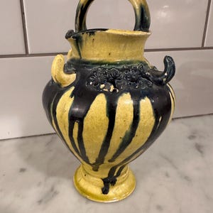 May include: A handmade ceramic vase with a yellow base and body, adorned with dark blue dripped paint. The vase features a curved handle, two side handles, and a decorative floral pattern near the top. The vase is approximately 10 inches tall.