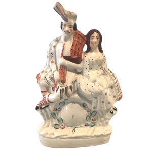 May include: A ceramic figurine of a couple in traditional Scottish attire. The man is wearing a kilt and playing a bagpipe, while the woman is wearing a dress with a plaid pattern. The figurine is painted in shades of white, red, blue, and green.
