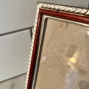 Puede incluir: Primer plano de un marco de fotos rectangular con un borde plateado y blanco en forma de cuerda. El marco tiene un borde interior rojo oscuro y un borde interior plateado. El marco est&aacute; en &aacute;ngulo, mostrando un vistazo de la foto en el interior.