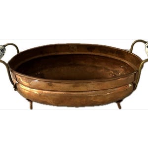 May include: A copper-colored, round serving bowl with a decorative rim and three small feet. The bowl has two handles with white ceramic grips featuring a blue floral design. A vintage kitchenware item.