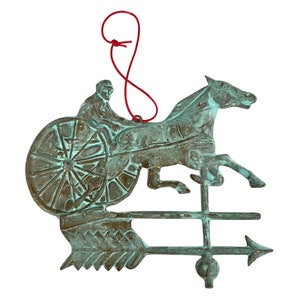Puede incluir: Un veleta de metal verde con un caballo y un jinete, con una cuerda roja para colgar. El caballo está al galope y el jinete sujeta las riendas. La veleta tiene una flecha direccional.