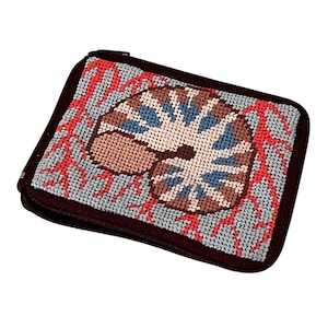 May include: A rectangular, zippered wallet with a cross-stitch design. The design features a tan, blue, and brown seashell-like pattern on a light blue background, with red accents. The wallet has a dark brown border and zipper.