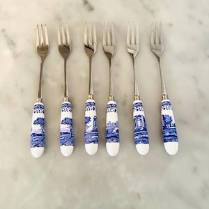 May include: A set of six silver-toned metal forks featuring white ceramic handles decorated with a blue and white landscape pattern. Each fork has three tines and a slender neck connecting the metal head to the cylindrical patterned handle.