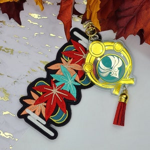 May include: A decorative keychain featuring an embroidered patch with red, orange, and teal maple leaf designs on a black background, attached to a gold-toned acrylic charm with a teal circular emblem and a small red fabric tassel.