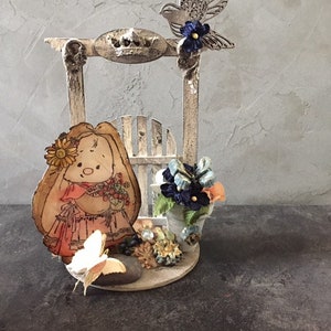May include: A white wooden frame with a decorative crown on top, a small white picket fence, and a paper cutout of a girl with brown hair holding a bouquet of flowers. A white butterfly is on the ground next to a gray stone. A blue bird with a blue flower is perched on the top of the frame.