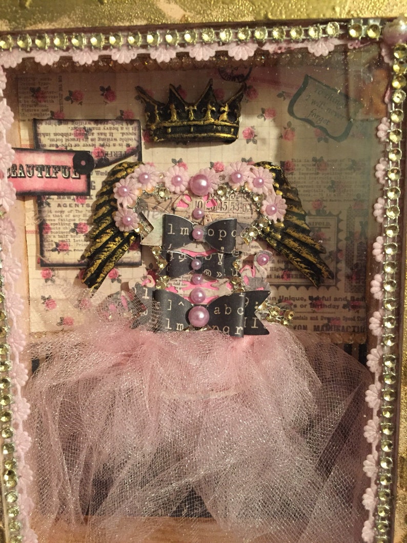 May include: A pink and gold shadow box with a crown, wings, and a tutu. The box has a pink tulle skirt and a black ribbon with the letters "1.m.o.p.o.  !  1  ]^  abc  1  m  agr" on it. The box also has a pink and white floral pattern.