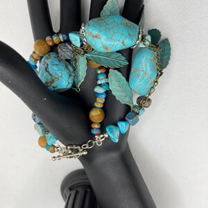 May include: A turquoise and brown beaded necklace with silver and gold accents. The necklace features large turquoise stones, smaller turquoise beads, and brown wooden beads. The necklace also has silver and gold leaf charms.