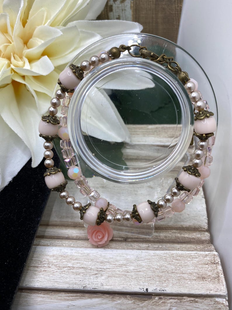 May include: A layered bracelet with pink beads, small pearls, and decorative metal details. A small pink rose charm hangs from the bracelet. The bracelet is displayed on a clear glass surface.