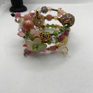 May include: A multi-strand bracelet with pink, green, and gold beads. The bracelet features a variety of beads, including flowers, crystals, and metal accents.