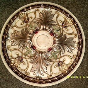 May include: A decorative ceiling medallion with a gold and silver leaf design. The medallion has a central circle with a raised edge and a floral pattern around the edge.