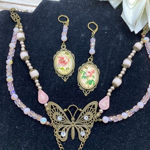 May include: A vintage-style jewellery set featuring a necklace and earrings. The necklace has a butterfly pendant with clear rhinestones and a chain. The earrings and pendant feature oval-shaped floral designs in a bronze setting, with pink and iridescent beads.