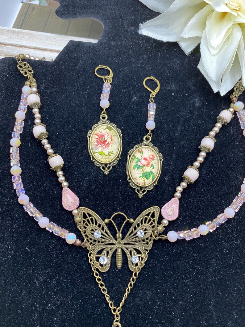 May include: A vintage-style jewellery set featuring a necklace and earrings. The necklace has a butterfly pendant with clear rhinestones and a chain. The earrings and pendant feature oval-shaped floral designs in a bronze setting, with pink and iridescent beads.