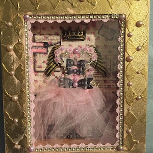 May include: A framed shadow box with a pink tulle tutu, gold wings, and a crown. The background is a collage of vintage paper with the words "I'm so happy" and "I'm a girl".