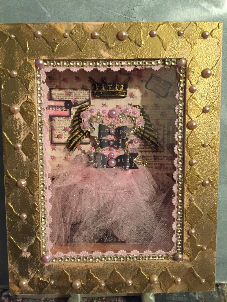 May include: A framed shadow box with a pink tulle tutu, gold wings, and a crown. The background is a collage of vintage paper with the words "I'm so happy" and "I'm a girl".