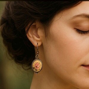May include: Close-up of a vintage-style earring. The earring features an oval pendant with a floral design in pink and yellow, set in a bronze-coloured metal frame. The earring is hanging from a hook.