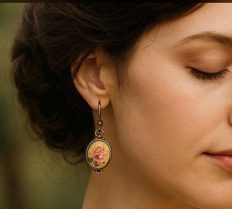 May include: Close-up of a vintage-style earring. The earring features an oval pendant with a floral design in pink and yellow, set in a bronze-coloured metal frame. The earring is hanging from a hook.