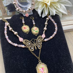 May include: A vintage jewellery set comprising a butterfly pendant necklace, earrings, and a bracelet. The necklace features a butterfly charm and an oval pendant with a floral design. The earrings and bracelet also have floral designs. The set is in shades of pink and gold.